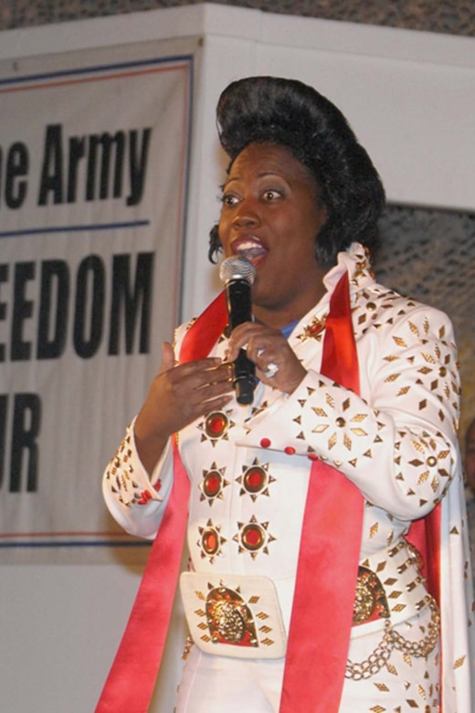 A woman in an Elvis costume speaks while servicemembers laugh in the background.