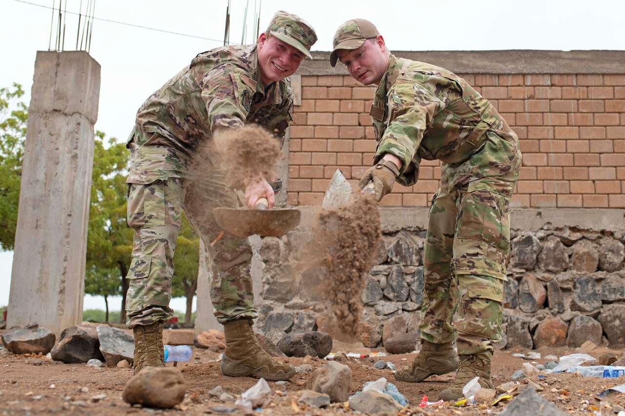 Two airmen shovel dirt next to each other while one smiles and looks at the camera.