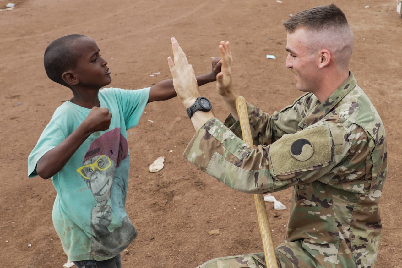A soldier kneels and holds up his hands as a child punches one of them.