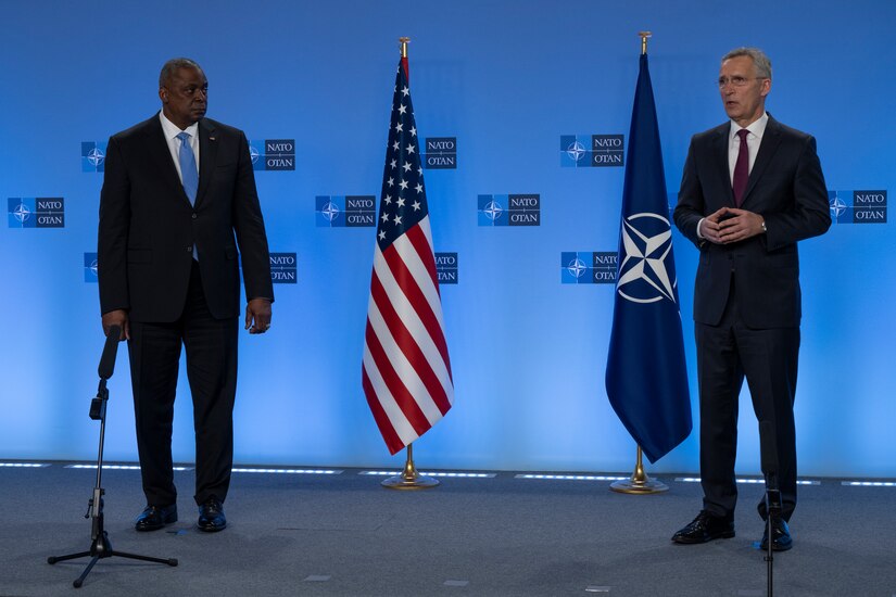 A man speaks while another listens while in front of a U.S. and NATO flags.