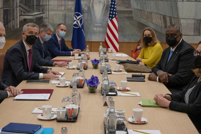 People sit at a conference table in front of a U.S. and NATO flags.
