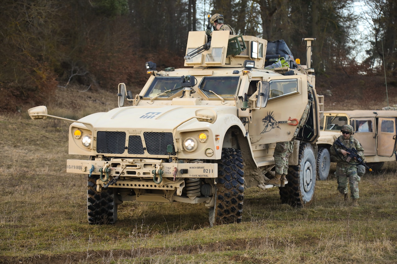 Soldiers walk around and sit atop a large-wheeled combat vehicle.