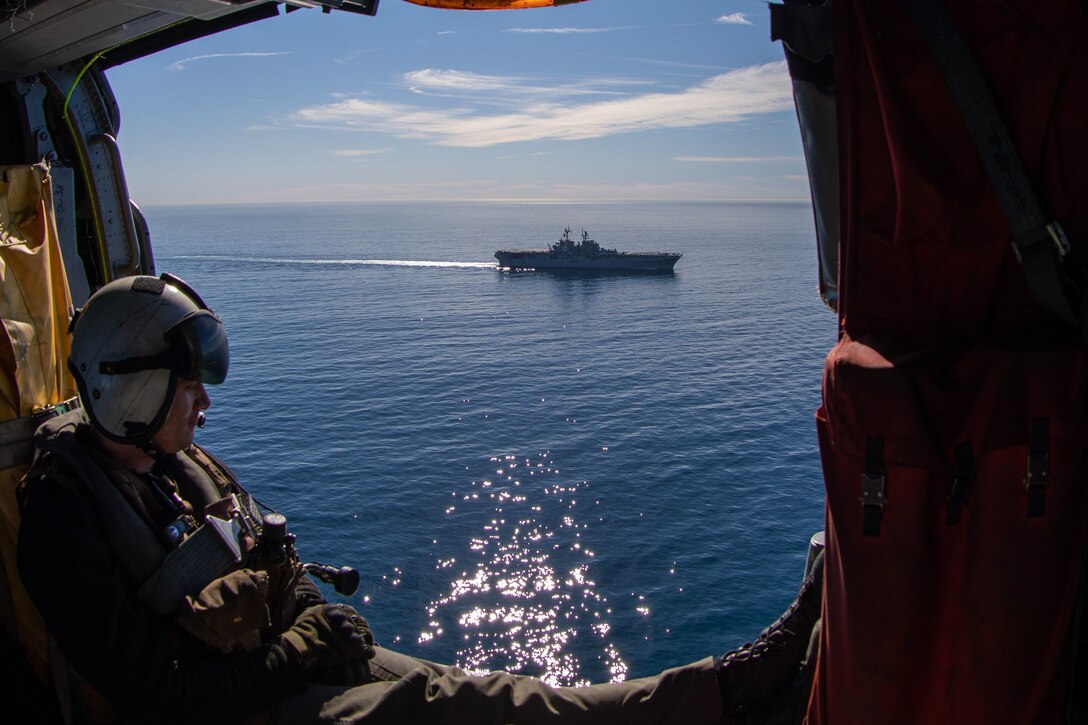 A sailor looks out from  a helicopter to a ship in the ocean.