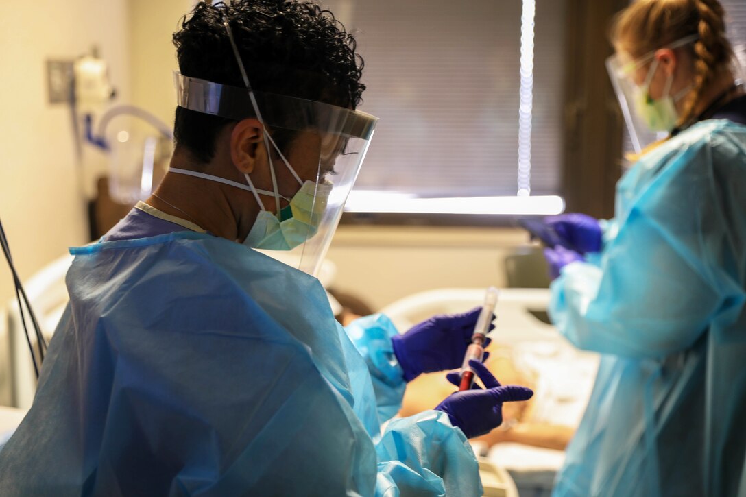 An airman wearing a face mask, gloves and gown cares works in a room with a civilian nurse.