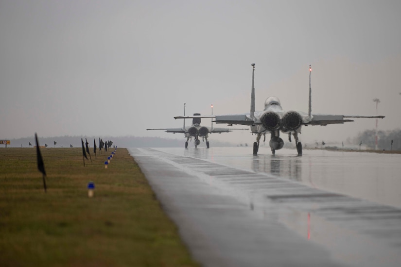 Two military jets taxi on a rainy runway.