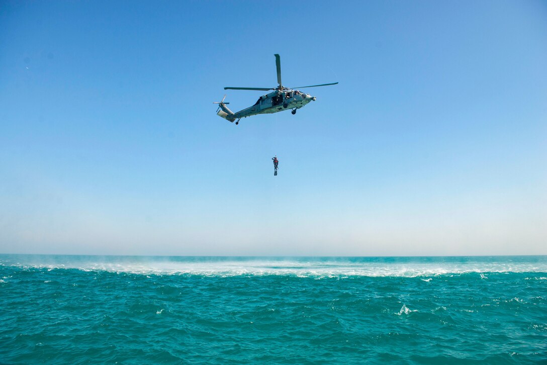 A service member holds on to a rope attached to a helicopter hovering over a body of water.