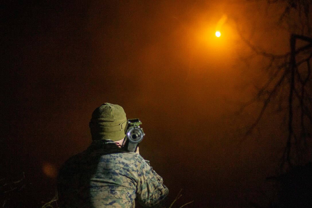 A Marine fires a weapon into a dark sky illuminated by an orange light.