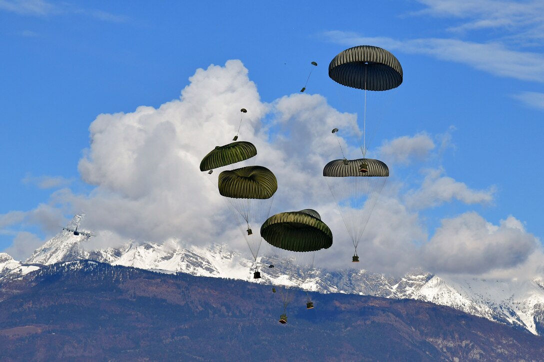 Packages attached to parachutes free fall near snowy mountains.