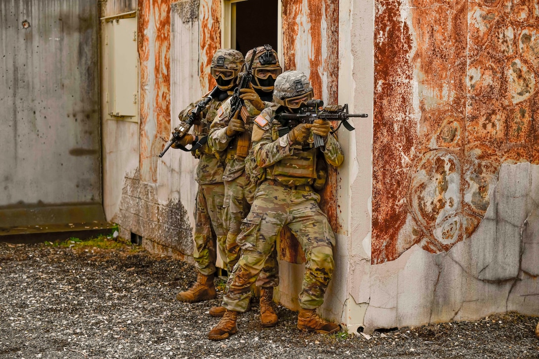 Three soldiers holding weapons stand next to a rusted a building during training.