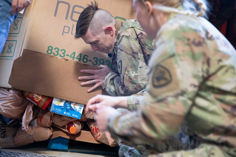 Soldiers put a box filled with packaged food on the ground of a warehouse.