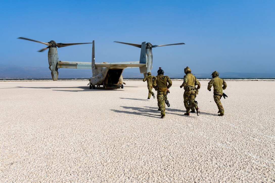 A group of airmen run toward an aircraft across sandy ground.