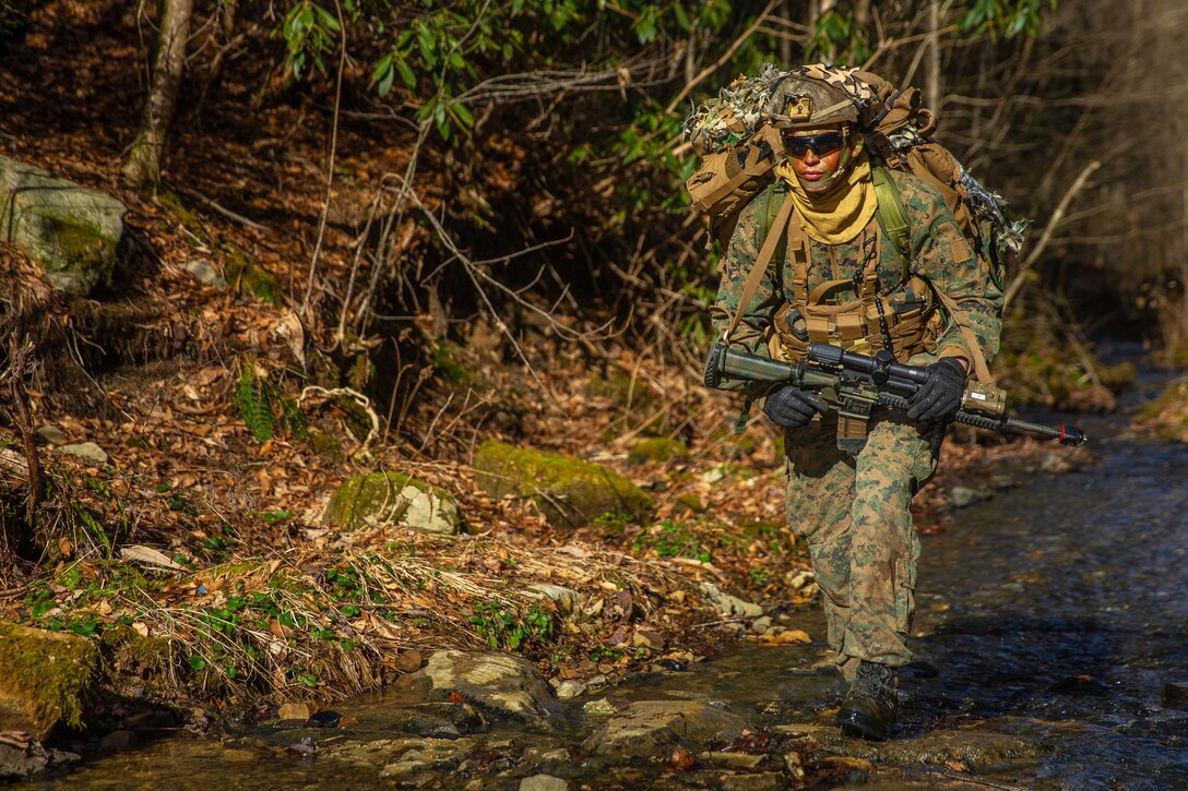 A Marine hikes across a creek in the woods.