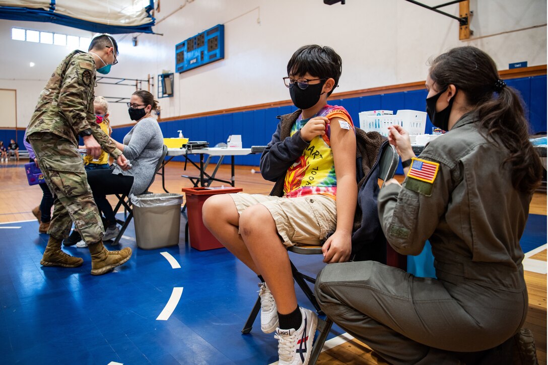 Two service members wearing face masks administers vaccines to two children.