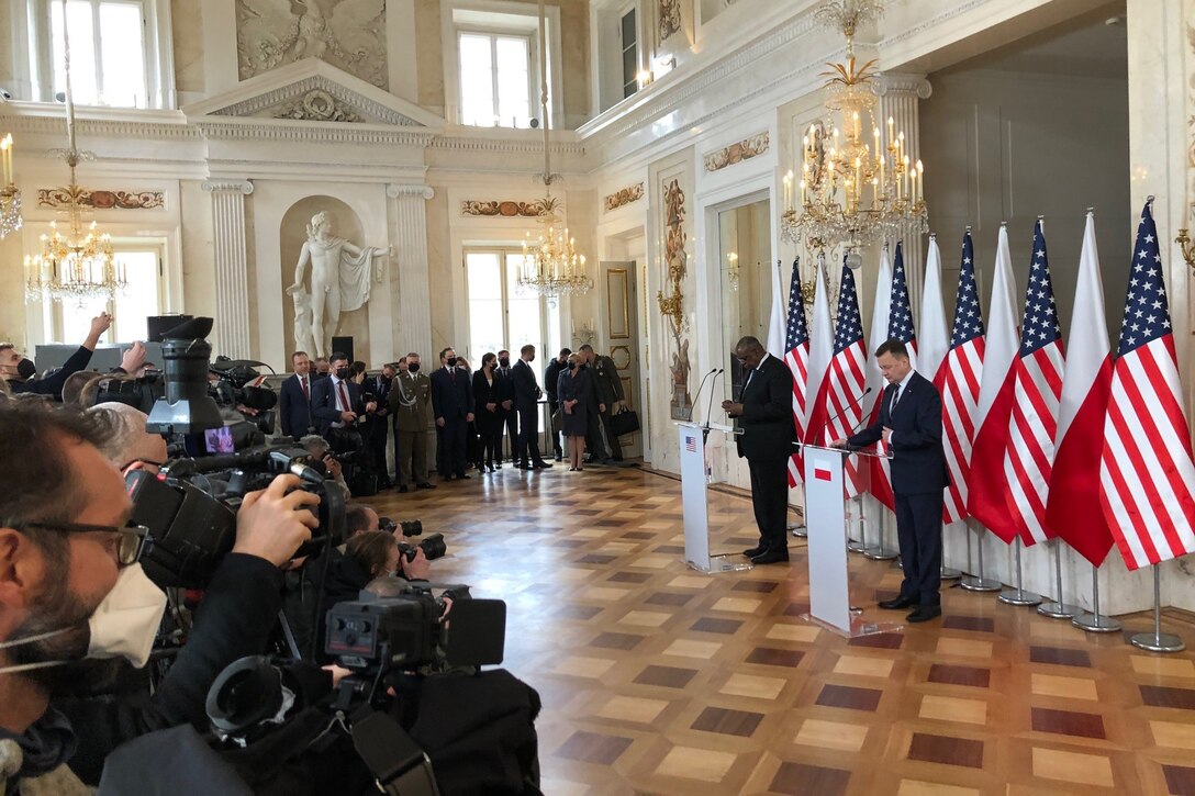 Two men stand at lecterns in front of a row of flags. Camera crews are in the foreground.