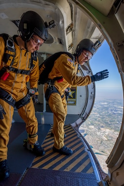Two Army Soldiers from the Army's parachute team wear yellow jumpsuits and stand in a plane's open door frame, preparing to jump.