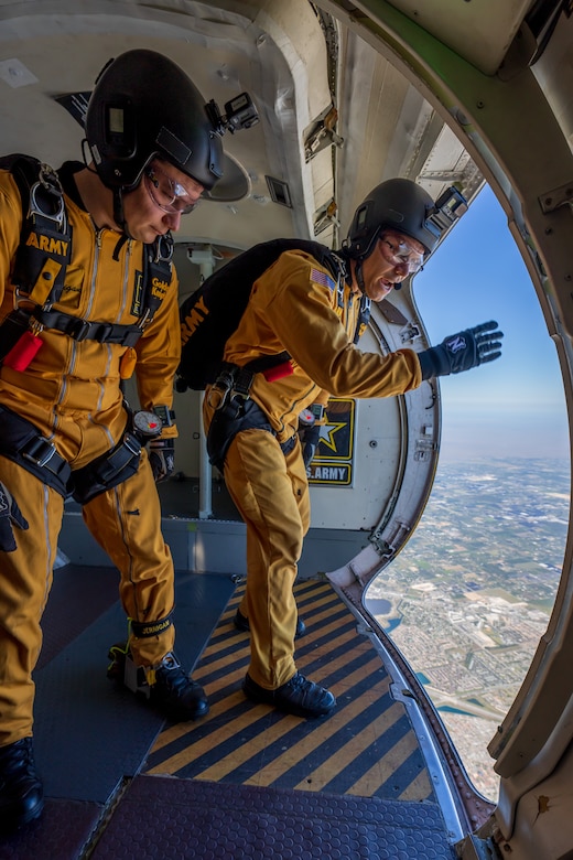 Two Army Soldiers in yellow jumpsuits stand in a plane's open door frame, preparing to parachute out.
