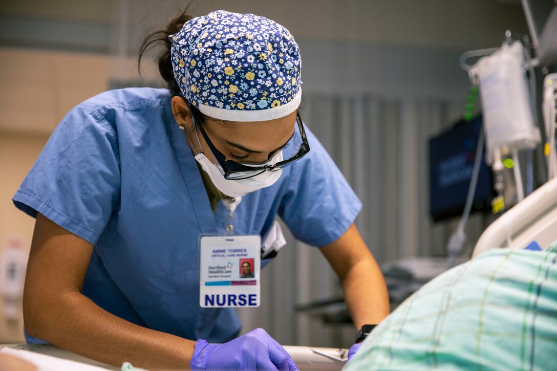 An airman wearing a face mask, gloves and surgical cap inserts an IV into a patient.