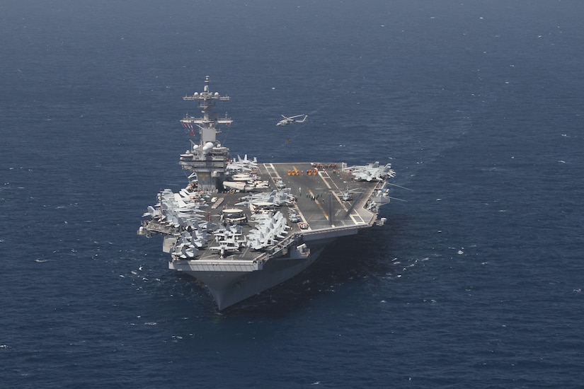 A large military ship moves through the ocean. Its deck is loaded with aircraft.