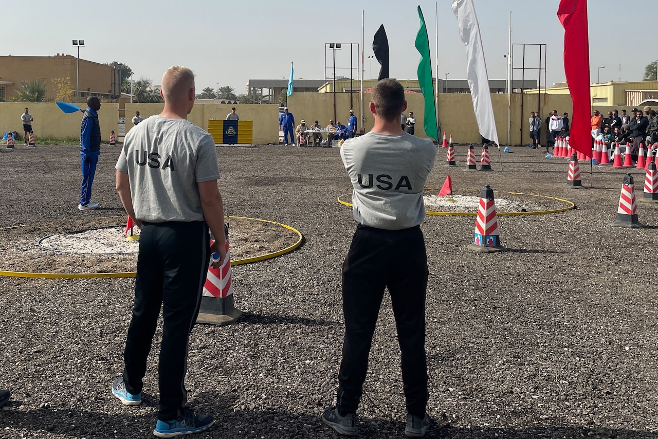 Two soldiers watch assembled athletes at an outdoor field marked by flags and cones.