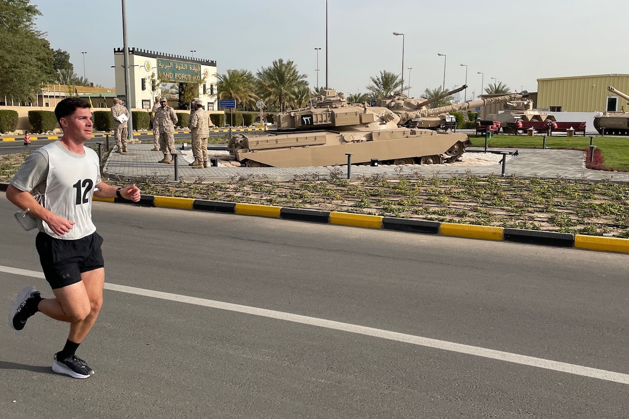 A soldier runs on asphalt past an outdoor tank display at a military base.