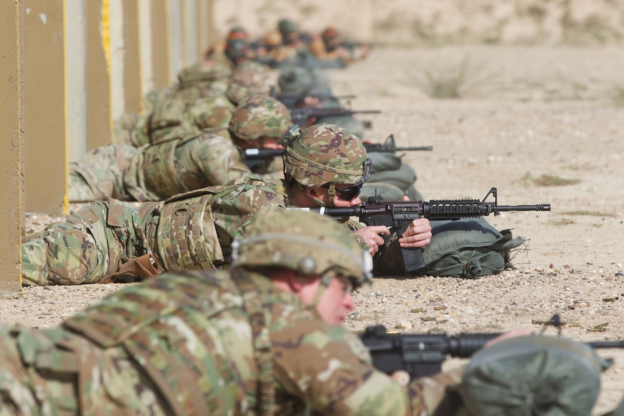 Soldiers fire weapons in the prone position at an outdoor range.