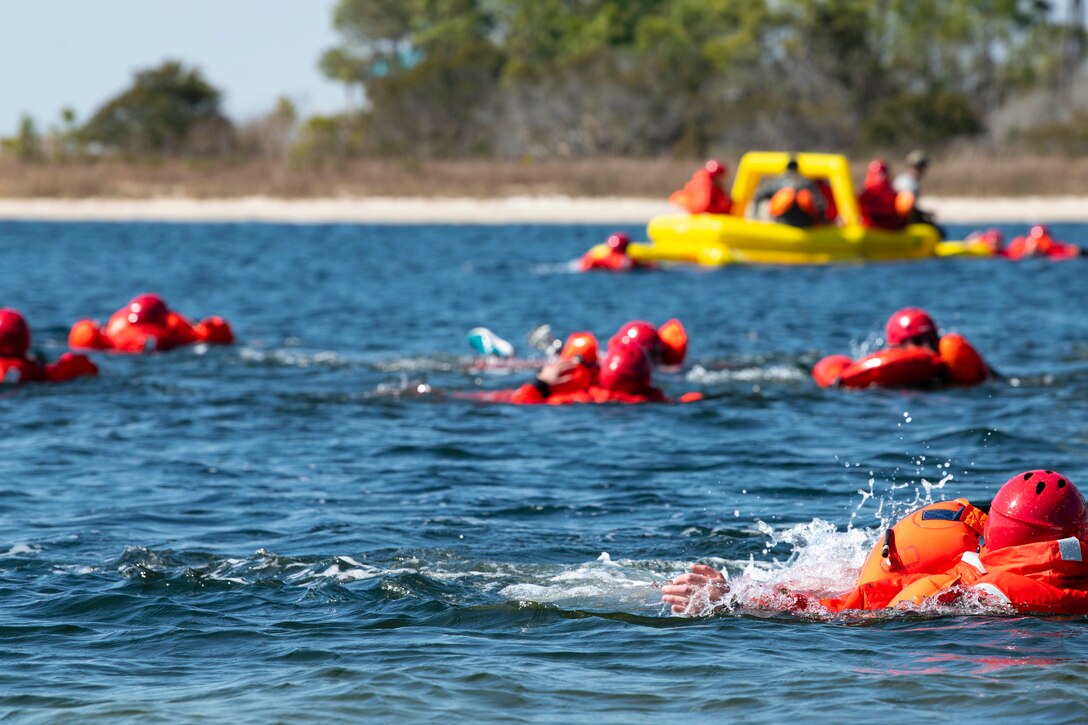 Airmen wearing red floating devices swim in a body of water near a small raft.