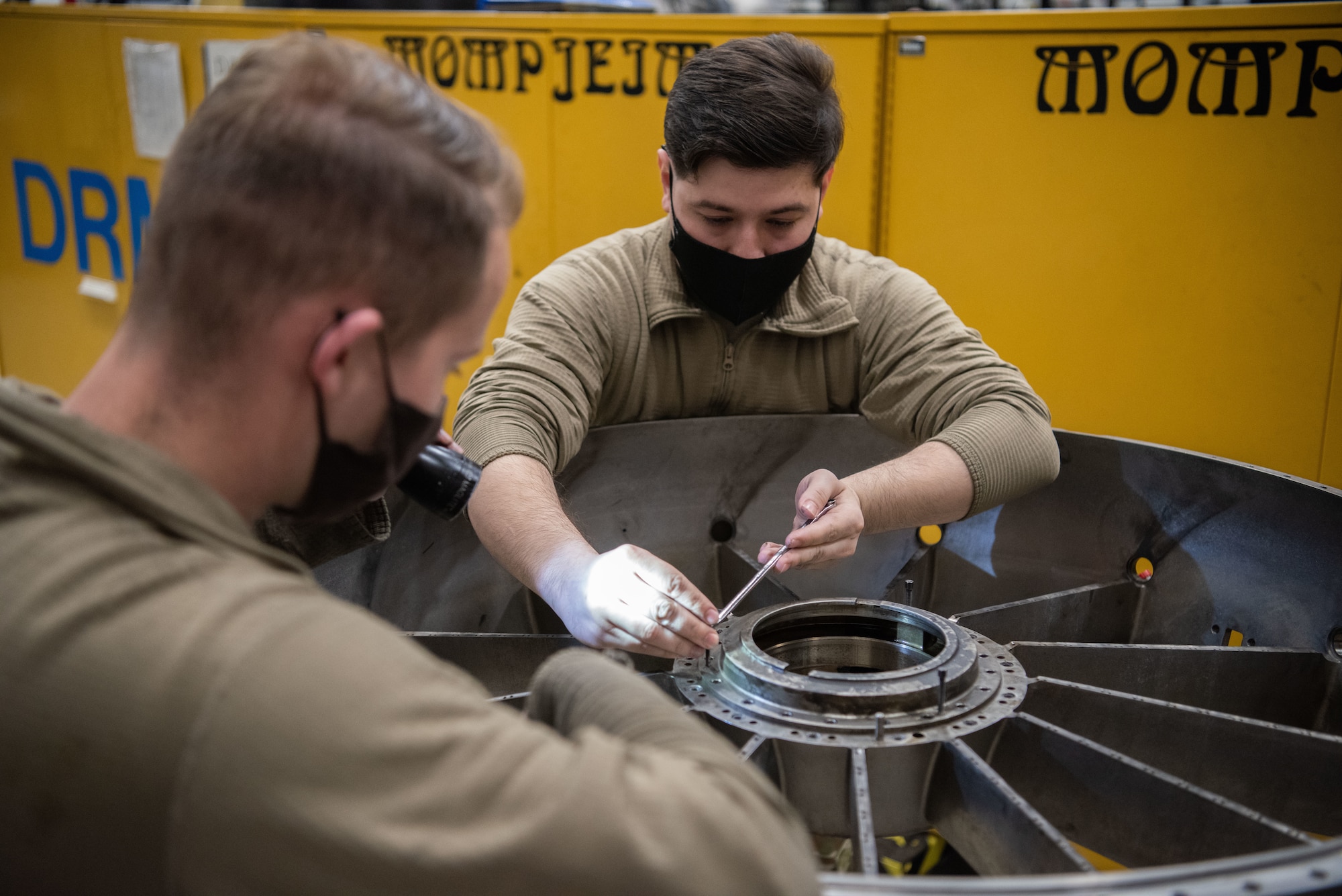 An Airman turns a screw on a jet engine piece while another Airman flashes a light on it.
