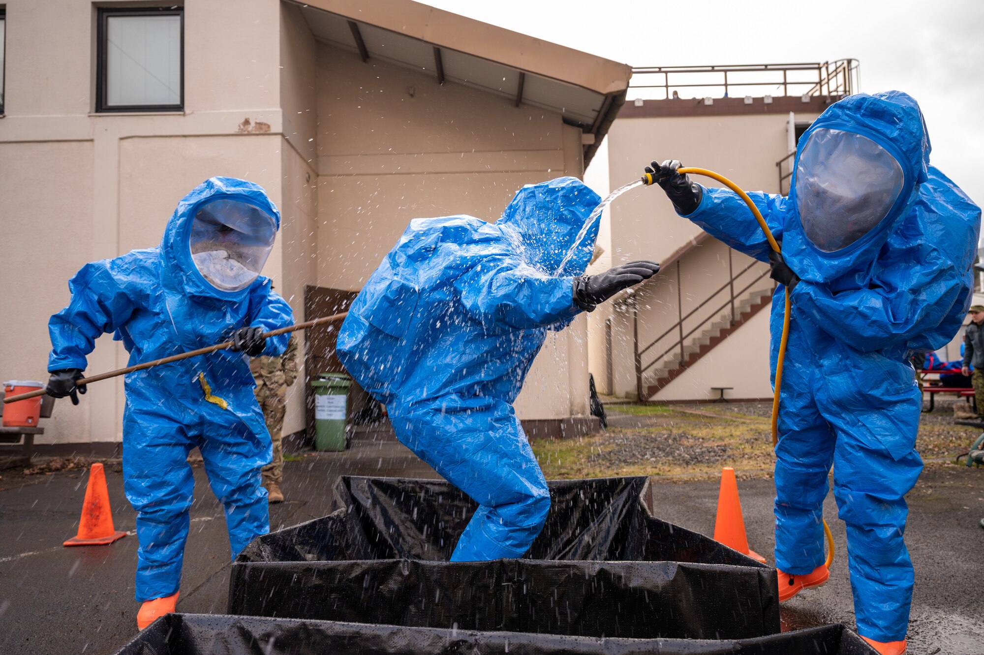 A person in a HAZMAT suit being sprayed with water and scrubbed with a brush by two other people in HAZMAT suits.