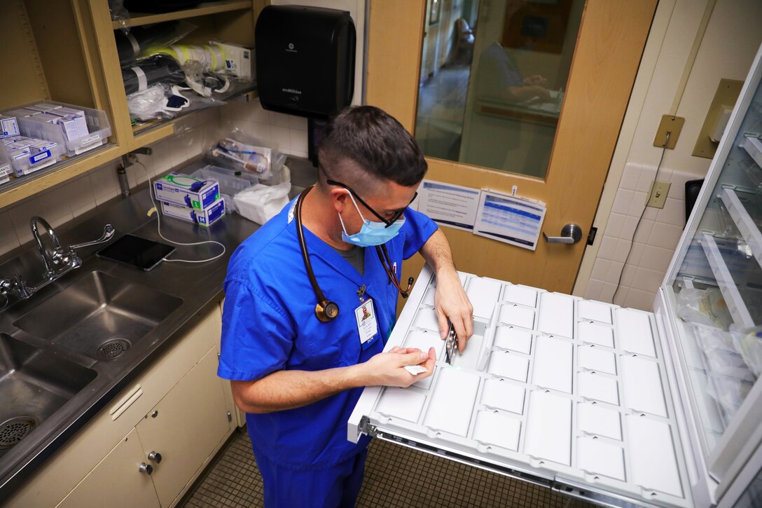 An Air Force airman wearing a face mask and a stethoscope takes out medication from a Omnicell cabinet.