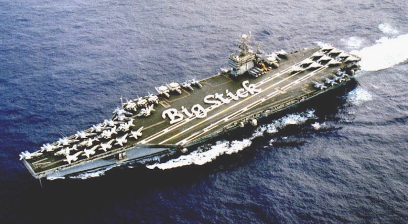 An aircraft carrier is underway with sailors in formation spelling “Big Stick.”