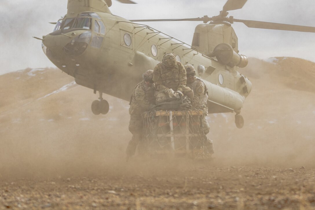 Soldiers hold onto a large container while a helicopter kicks up dust behind them.