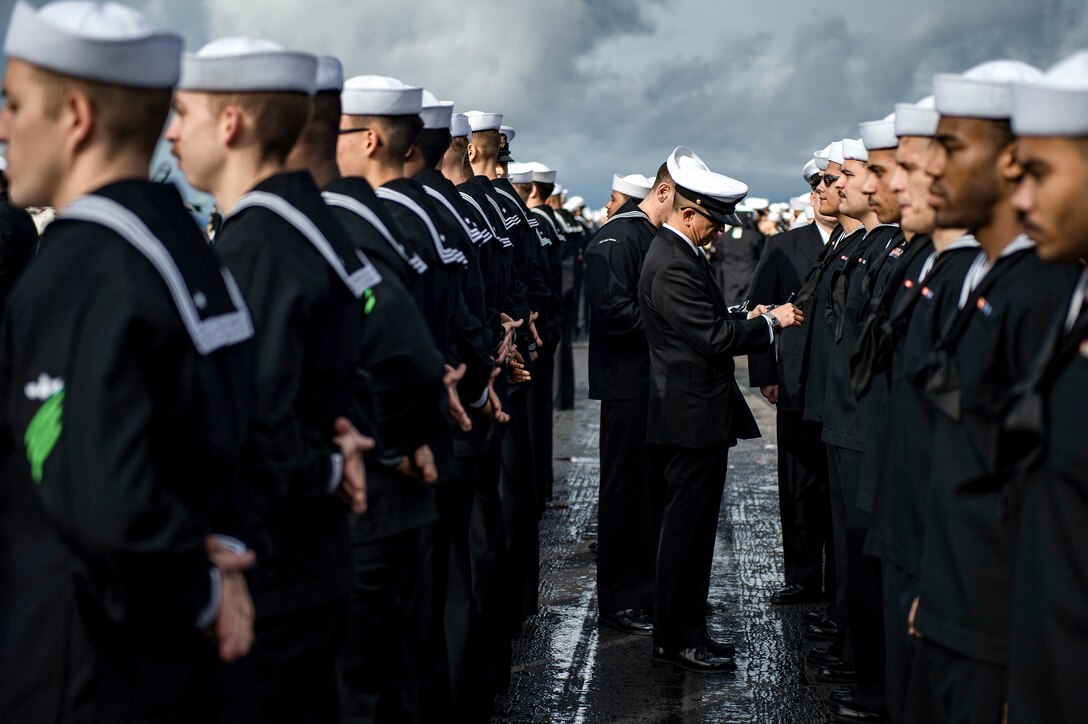 Sailors stand in rows wearing dress uniforms as a fellow sailor inspects them.