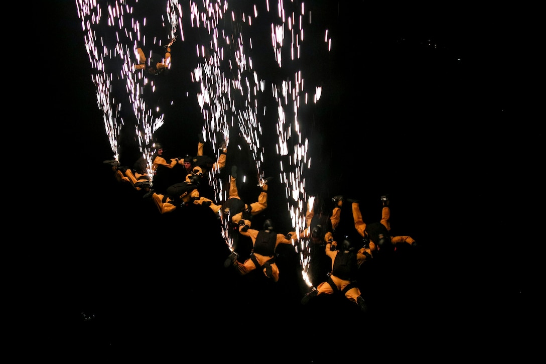 Flares shoot up from a group of Army parachutists descending in formation in a dark sky.