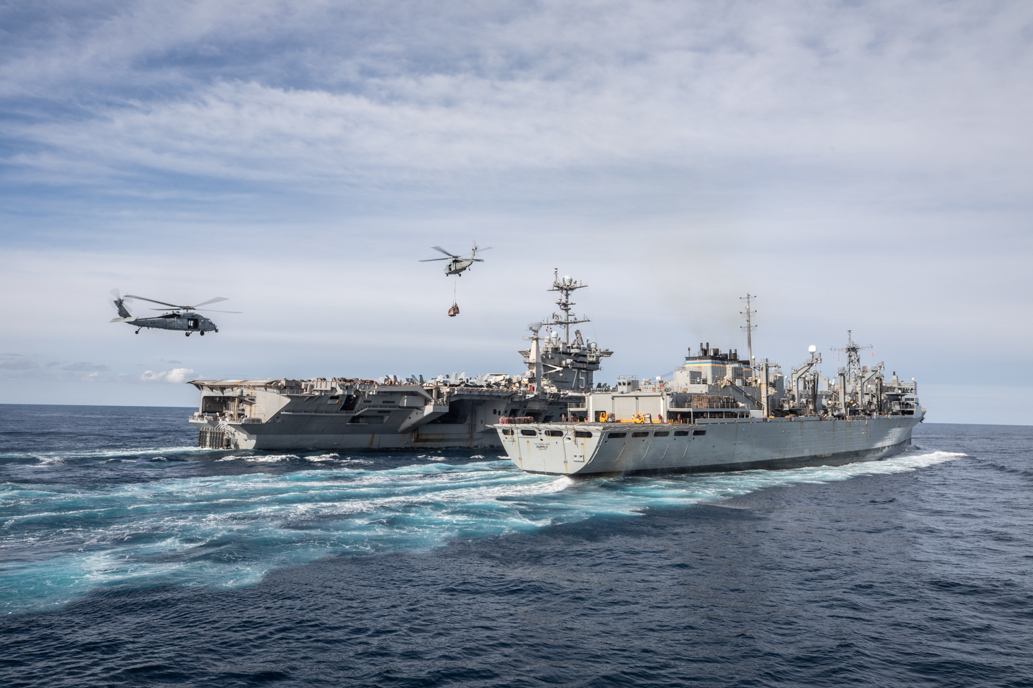 U.S. Navy Nimitz-class aircraft carrier USS Harry S. Truman (CVN 75), left, conducts a replenishment-at-sea with U.S. Navy Supply-class fast combat support ship USNS Supply (T-AOE-6), right. In the air, two MH-60S Sea Hawk helicopters assigned to the “Dragonslayers” of Helicopter Sea Combat Squadron (HSC) 11 conduct fly supplies between the two ships during vertical replenishment operations.