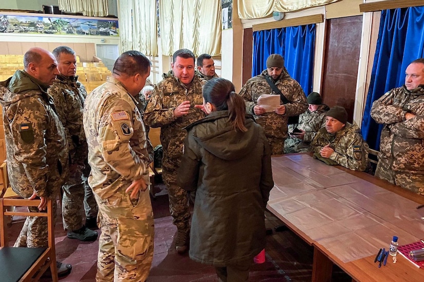 A foreign service member speaks to a woman wearing a jacket while several other domestic and foreign service members look on.