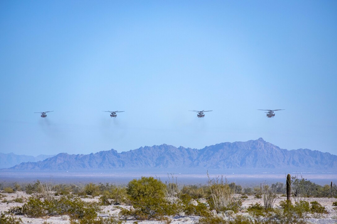 Four helicopters fly over a desert area with mountains in the background.