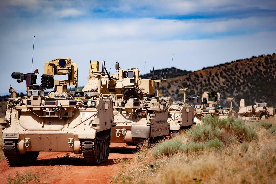 Tactical vehicles in single-line formation travel down a dirt road,