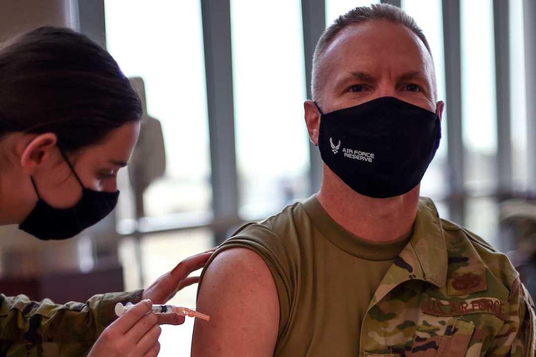 A medical tech wearing a face mask leans over with a syringe to administer a COVID-19 vaccine to a senior enlisted leader wearing a face mask while sitting.