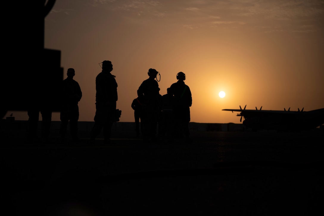 Airmen stand on a tarmac near an aircraft under a sunlit sky.