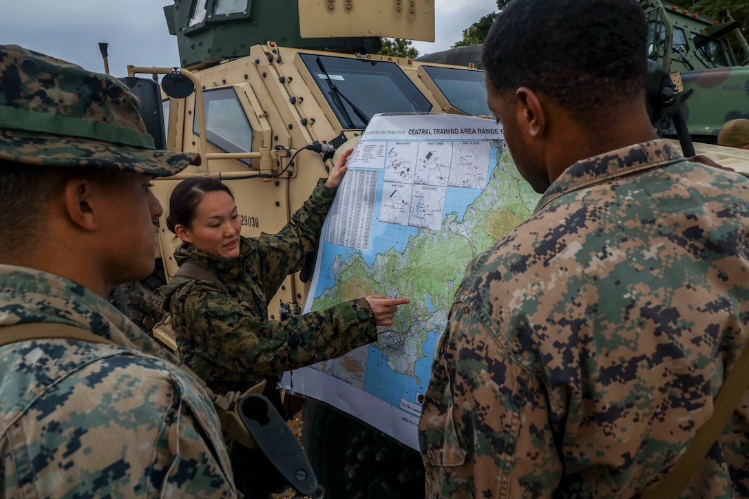 A Marine points to a spot on a map she's holding up against a military vehicle as two other Marines observe.