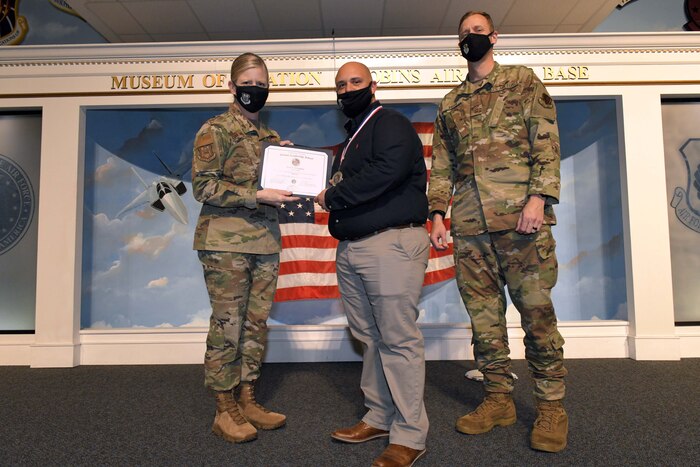 Photo shows individual standing with certificate between two other people.