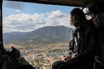 Soldier looks out helicopter doorframe over mountains and military base.