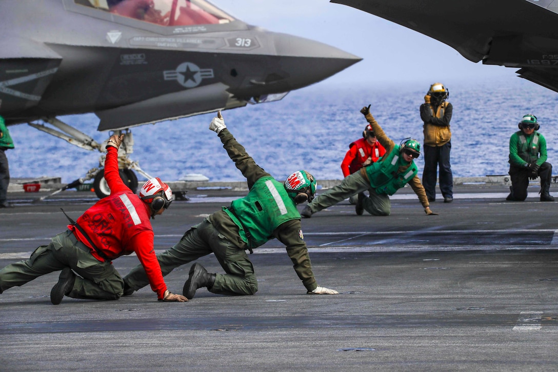 Three Marines kneel on the deck of a ship while signaling toward an aircraft.