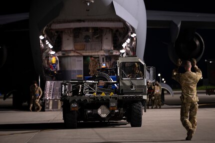A military aircraft sits on a runway.  Airmen load cargo aboard the aircraft.