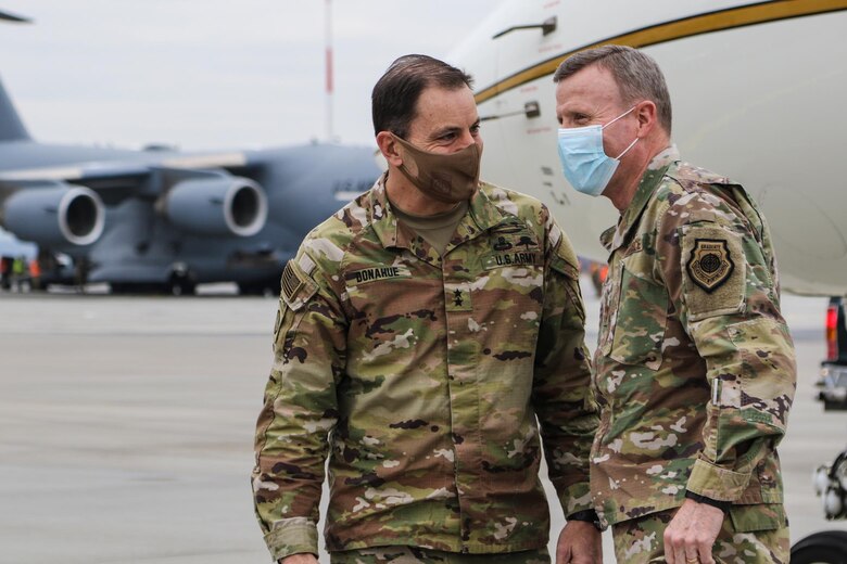 Two men wearing military uniforms talk; a large military plane is in the background.