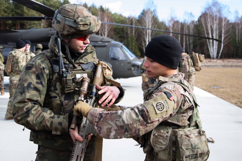 Two service members examine a weapon; a helicopter and other service members are in the background.