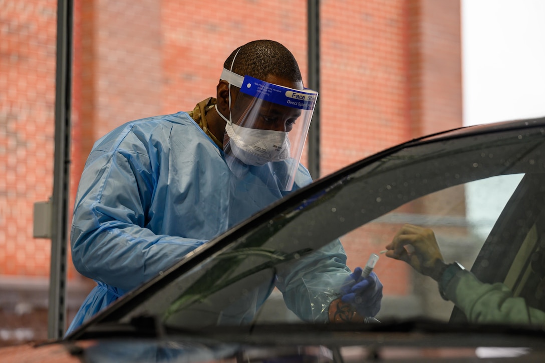 A soldier wearing personal protective equipment holds a vial while collecting a nasal swab from a patient.