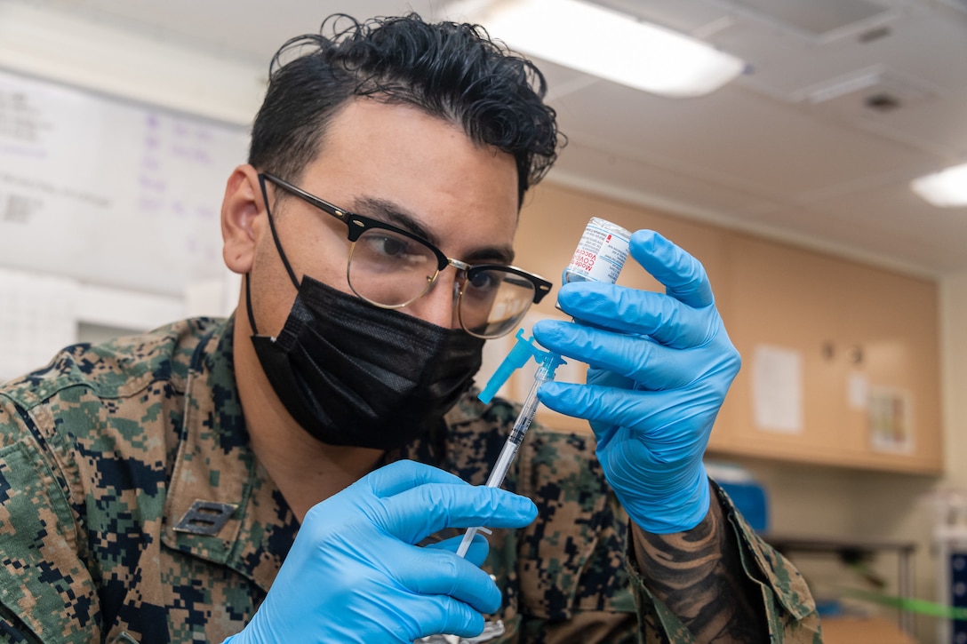 A sailor wearing a face mask and gloves holds a syringe while inserting the needle into a small bottle to prepare a COVID-19 vaccine booster.
