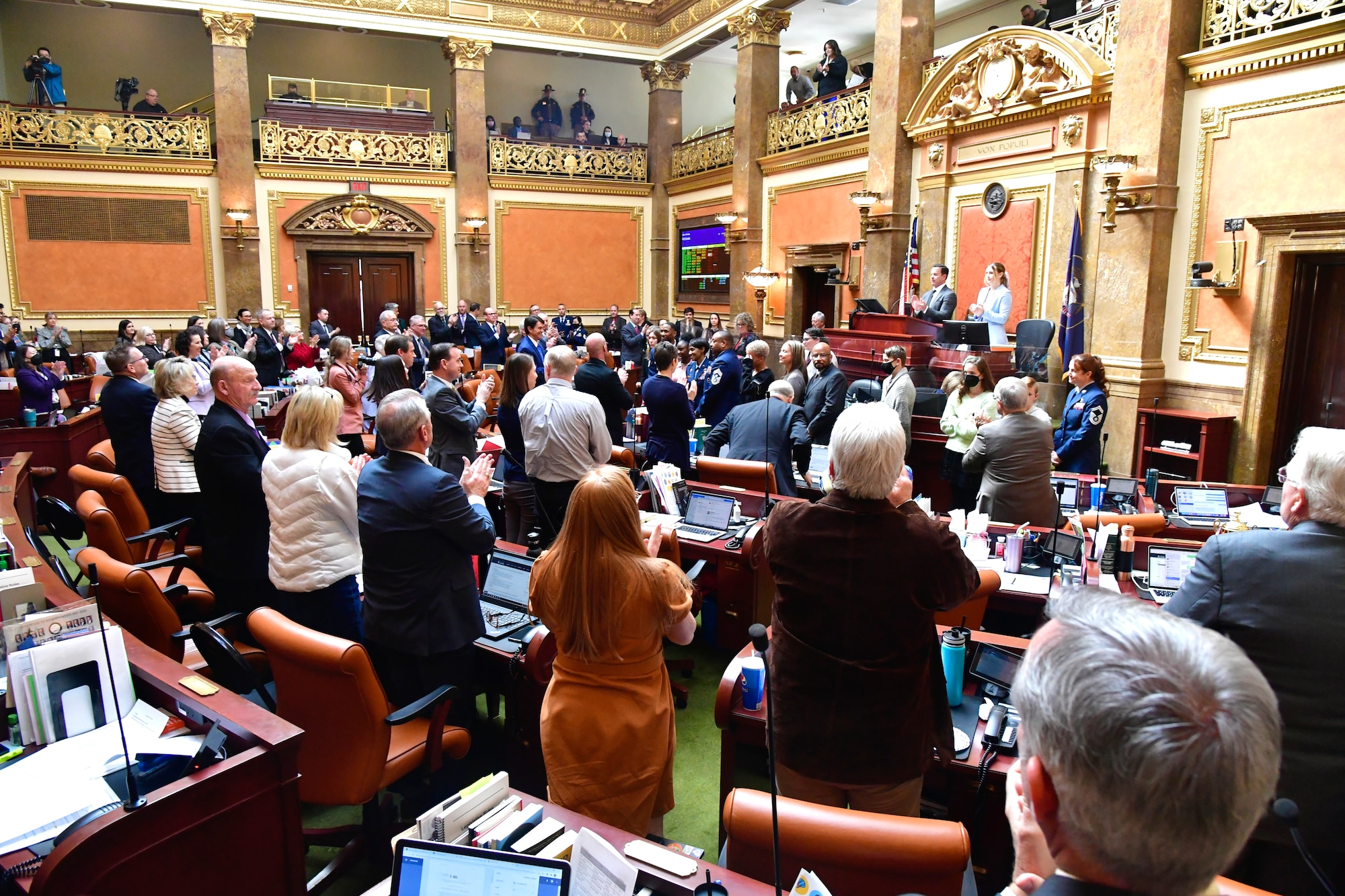 Military and civilian families stand in front of applauding legislators inside the chamber.