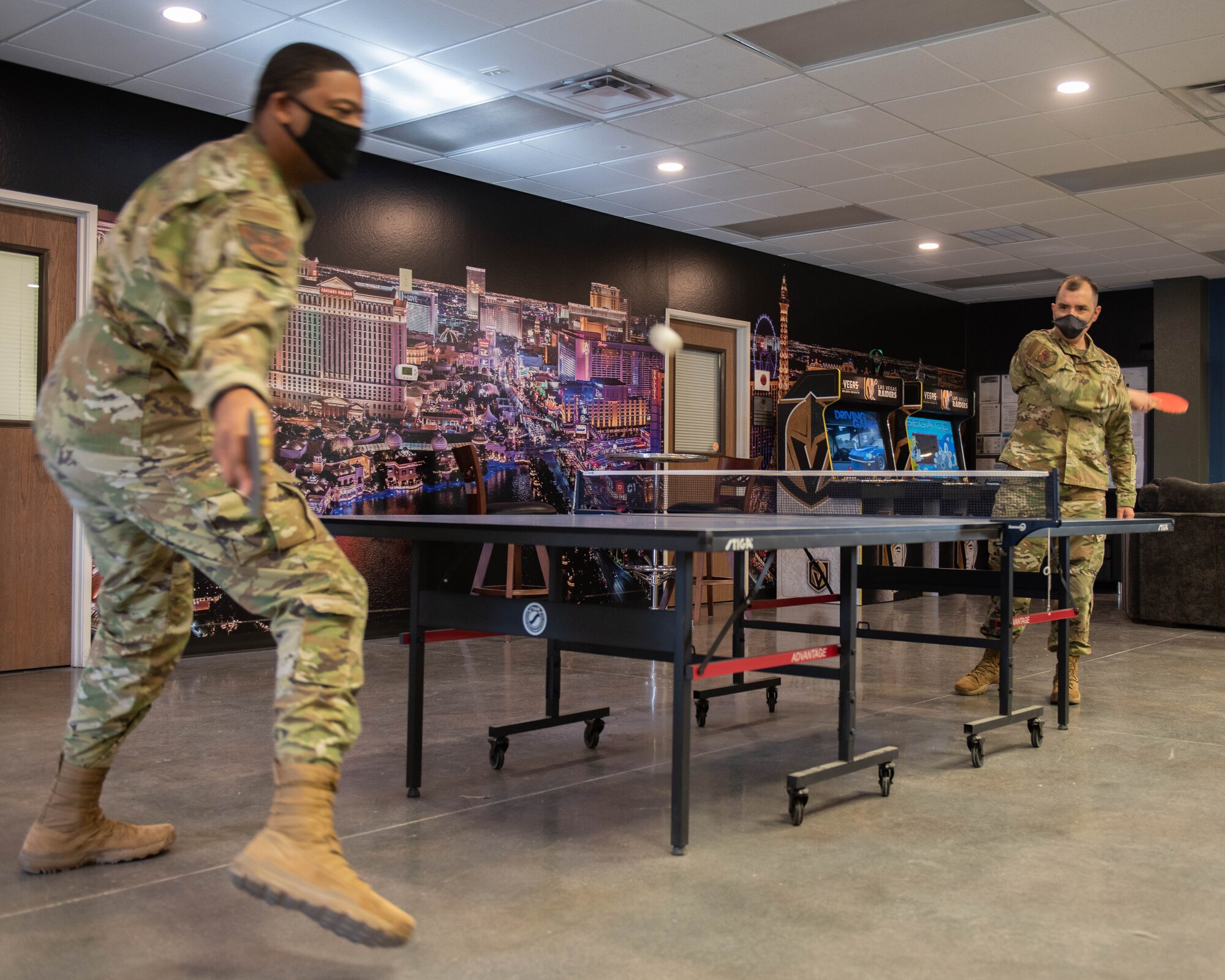 Capt. Taylor stands on the right side of the ping pong table. After hitting the ball, the ball flew to the side of his opponent's. His opponent, another male Airman, prepares himself to hit the ball back to Taylor. Behind Taylor, there two are arcade games and the wall is a wall-paper of the Las Vegas strip.